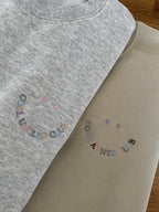 Zwei Sweater in beige und ash heather mit jeweils einer Stickerei, die wie ein Smiley aussieht: Cool Uncles Club und Cool Aunts Club.