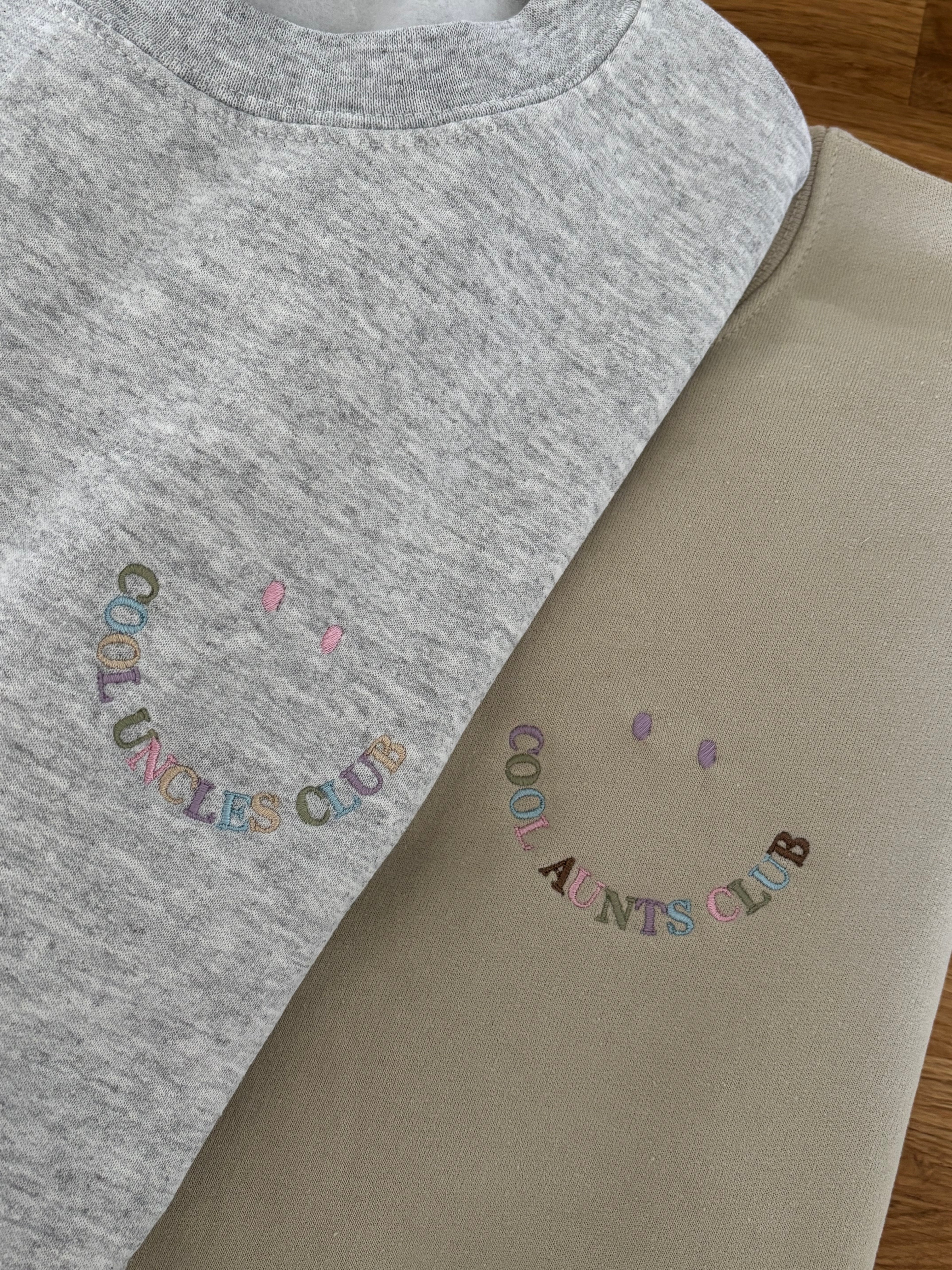 Zwei Sweater in beige und ash heather mit jeweils einer Stickerei, die wie ein Smiley aussieht: Cool Uncles Club und Cool Aunts Club.