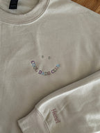 Ein beiger Sweater mit einer bunten Stickerei in Form eines Smileys: Cool Dads Club. Auf dem Ärmel ist der Name des Kindes gestickt "Henri".