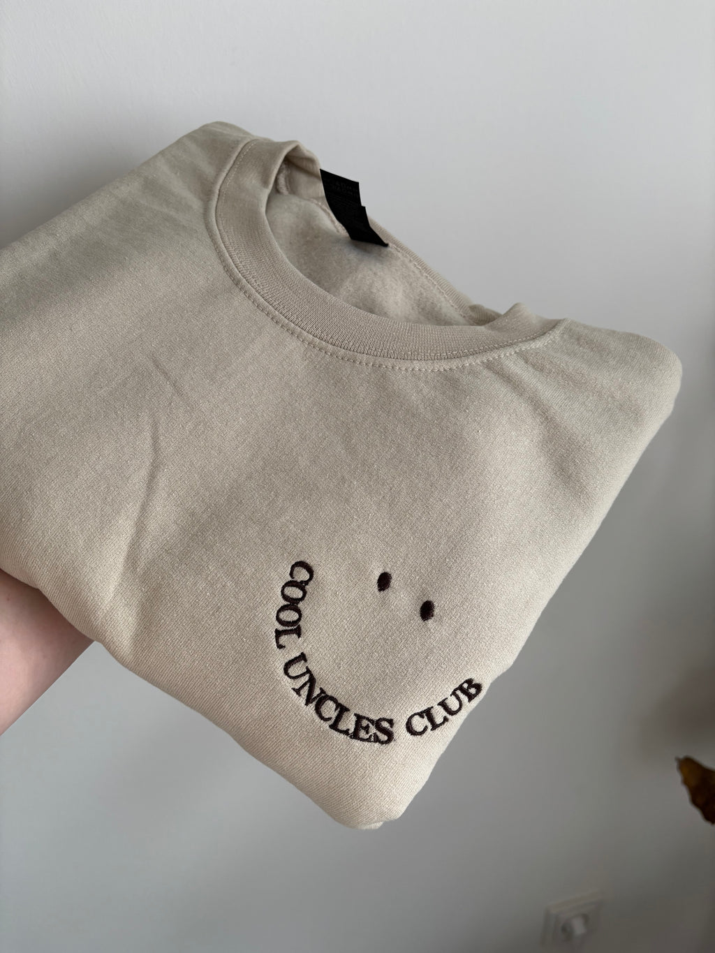 Ein beiger Sweater mit einer dunkel braunen Stickerei Cool Uncles Club.