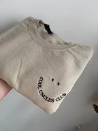 Ein beiger Sweater mit einer dunkel braunen Stickerei Cool Uncles Club.
