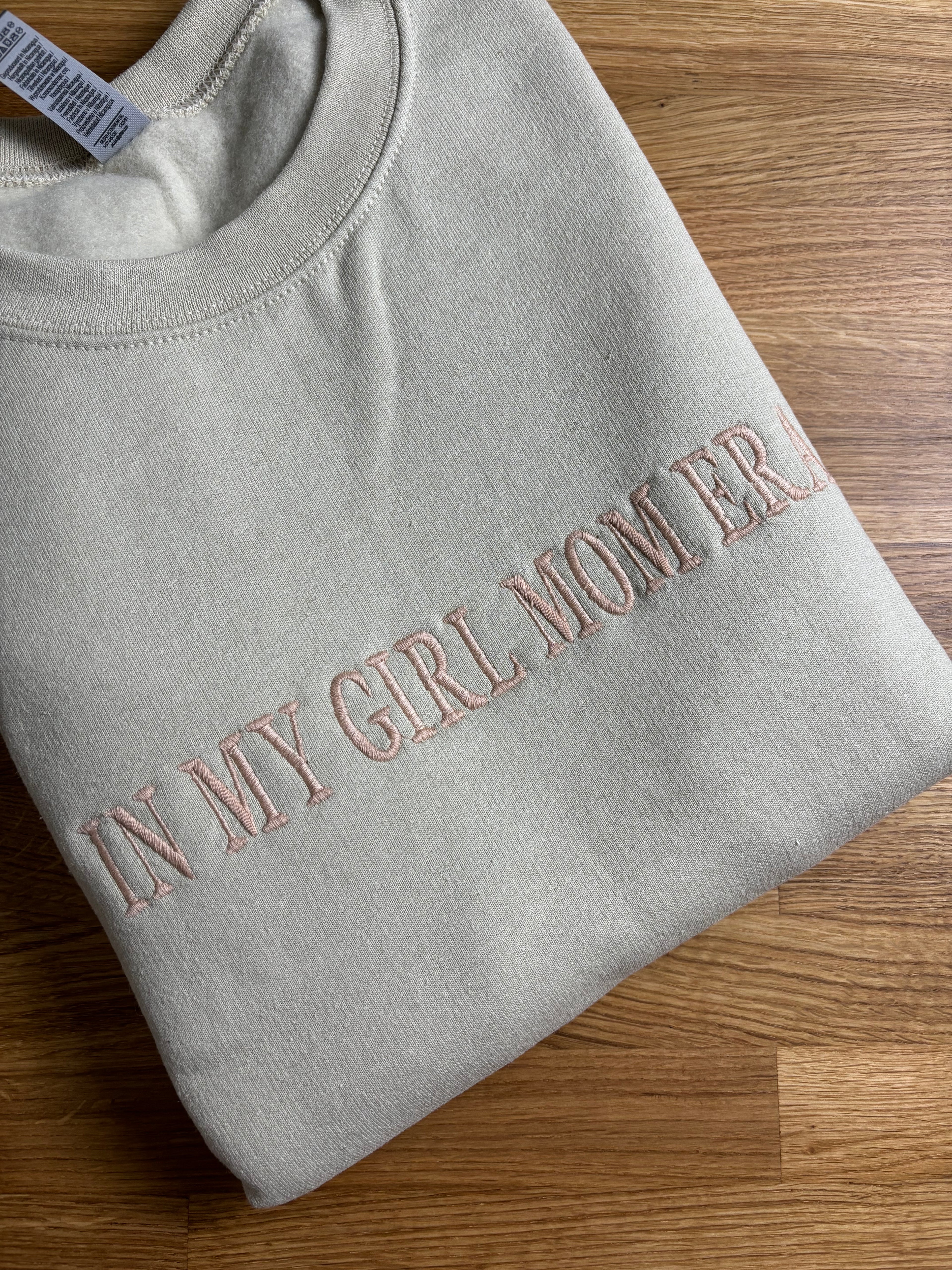 Ein Sweater in der Farbe Beige mit einem personalisierten Stick vorne auf der Brust mittig in poudre "In my girl mom era".