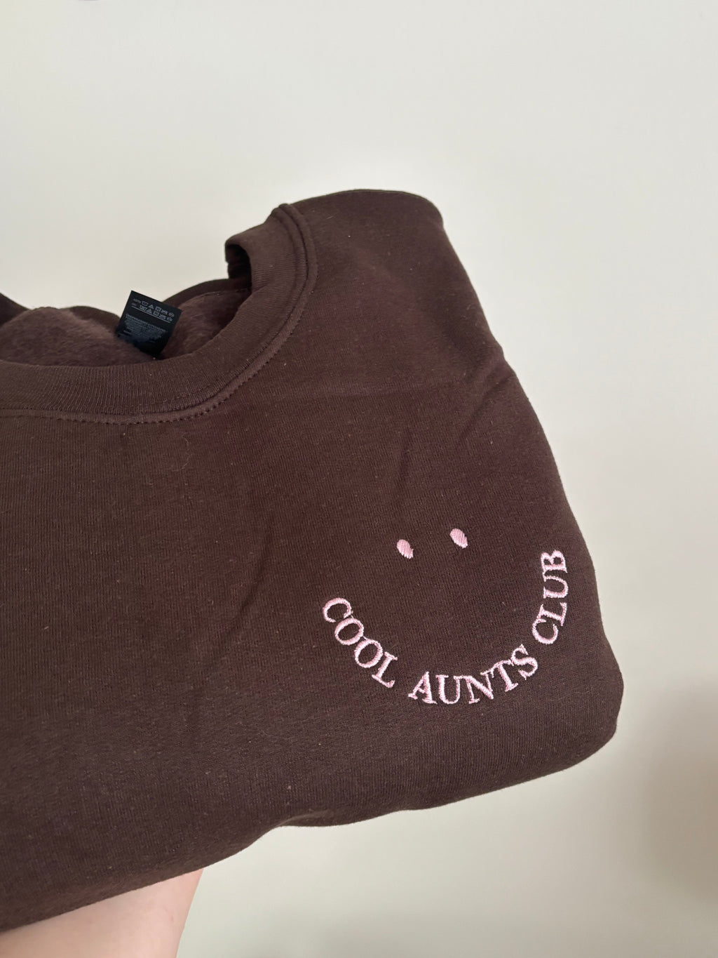Der Sweater in der Farbe Dark Chocolate ist mit einer Stickerei versehen: Cool Aunts Club. Die Stickerei ist in der Form eines Smileys.