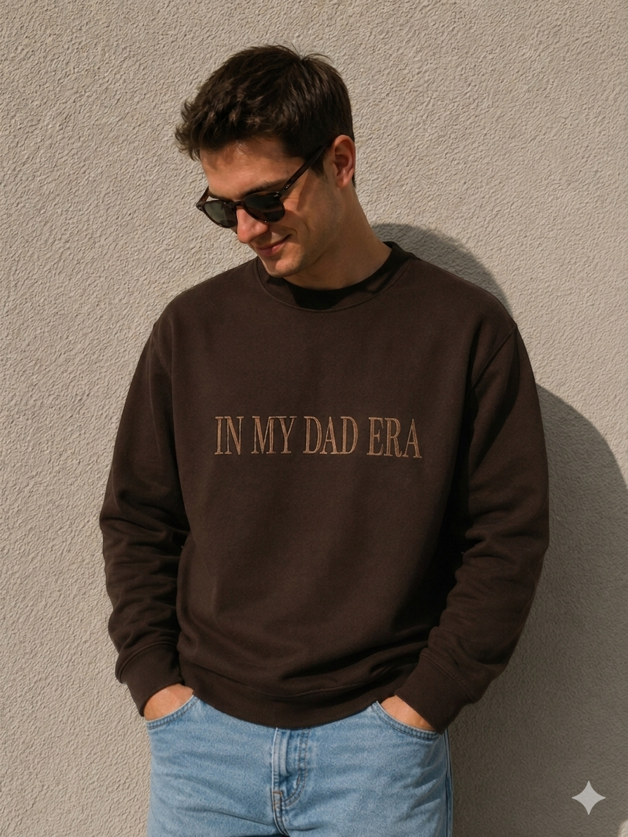 Ein junger Mann steht vor einer hellen, strukturierten Wand im Sonnenlicht. Er trägt eine Sonnenbrille, blickt lächelnd nach unten und hat die Hände in den Taschen seiner hellblauen Jeans. Er trägt einen dunkelbraunen Crewneck-Pullover, auf dem in gerader, beigefarbener Stickerei zentriert der Schriftzug „IN MY DAD ERA“ zu sehen ist.