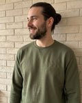 Ein Mann mit langen Haaren trägt einen Sweater in der Farbe Military Green. Der Sweater ist mit einer Stickerei versehen: DAD est. 2026.