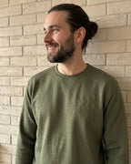Ein Mann mit langen Haaren trägt einen Sweater in der Farbe Military Green. Der Sweater ist mit einer Stickerei versehen: DAD est. 2026.