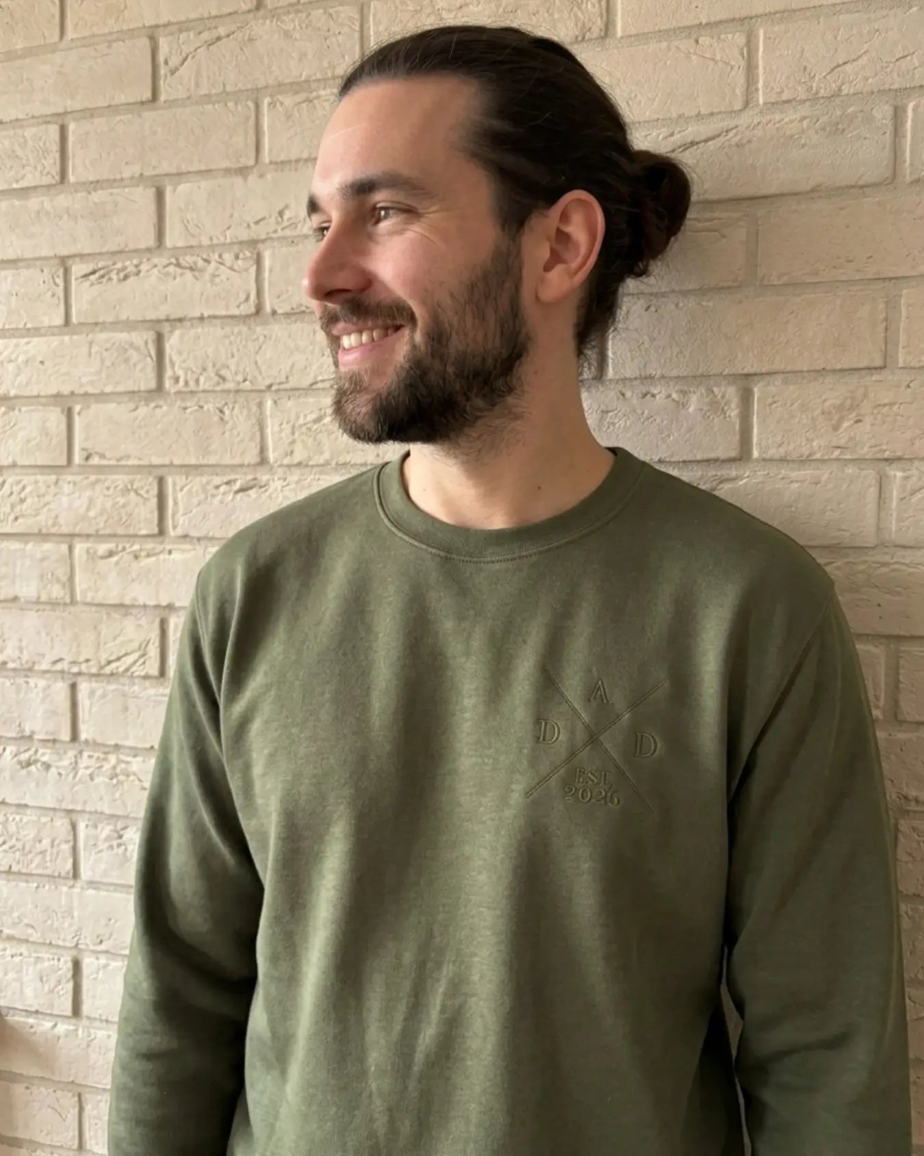 Ein Mann mit langen Haaren trägt einen Sweater in der Farbe Military Green. Der Sweater ist mit einer Stickerei versehen: DAD est. 2026.