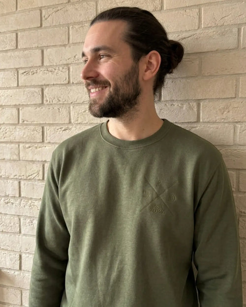 Ein Mann mit langen Haaren trägt einen Sweater in der Farbe Military Green. Der Sweater ist mit einer Stickerei versehen: DAD est. 2026.