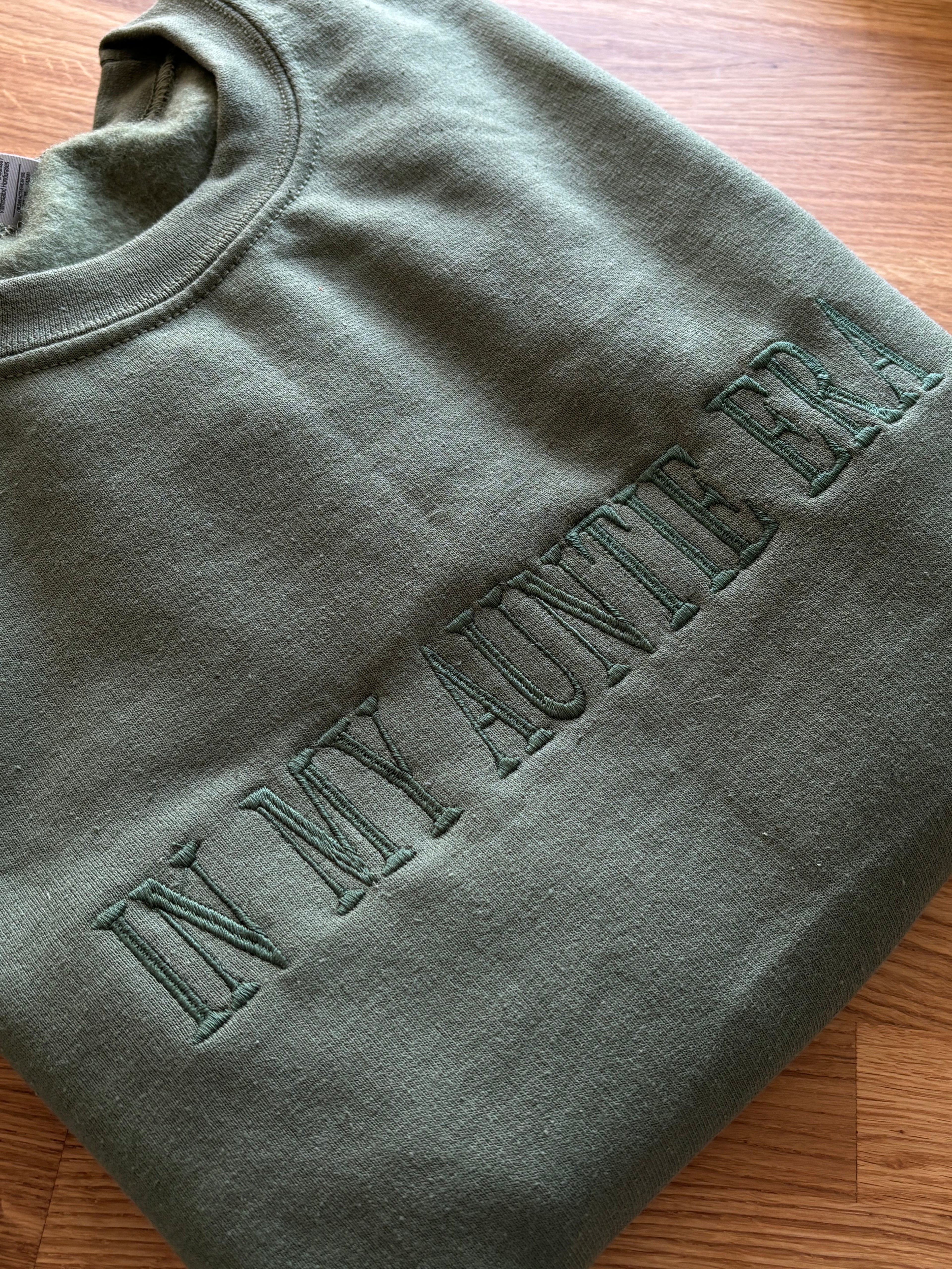Ein Sweater in der Farbe Military Green mit einem bestickten Wunschtext vorne auf der Brust mittig in green "In my Auntie era".
