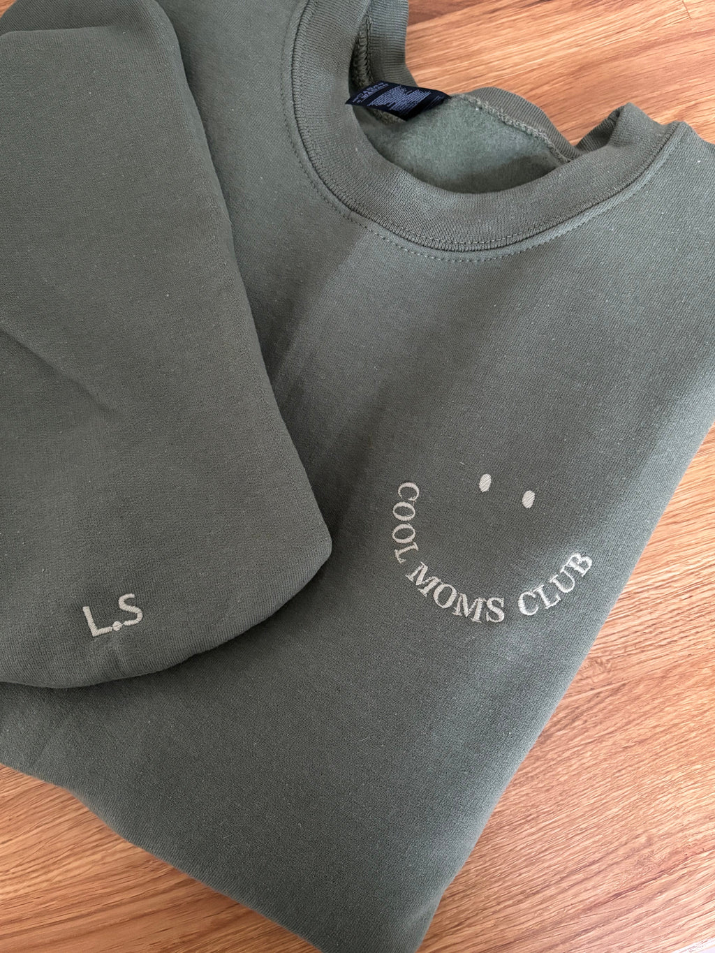 Ein gefaltetes grünes Sweatshirt liegt auf einem Holztisch. Auf der Brust ist ein lächelndes Gesicht aufgestickt, wobei der Mund aus dem Schriftzug "COOL MOMS CLUB" in grüner Farbe besteht und zwei Punkte darüber die Augen darstellen. Dazu gibt es den passenden Ärmelstick.