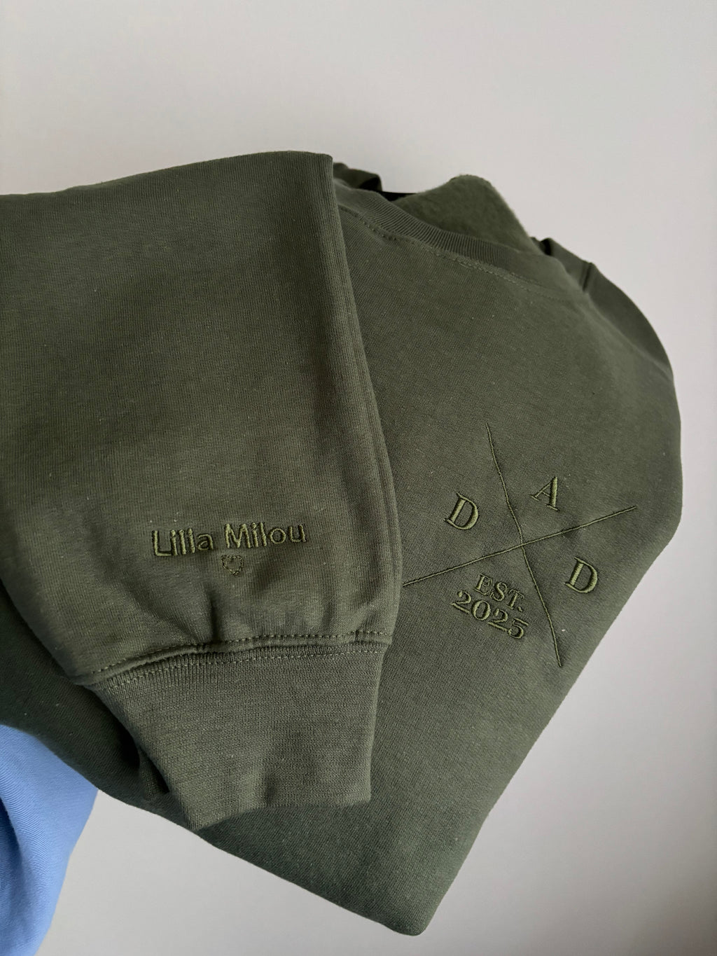Ein Sweater in der Farbe Military Green mit einem Koordinaten Kreuz auf der linken Brust gestickt. Im Kreuz steht Dad est. 2025. Auf dem Ärmel ist der Kindername gestickt.
