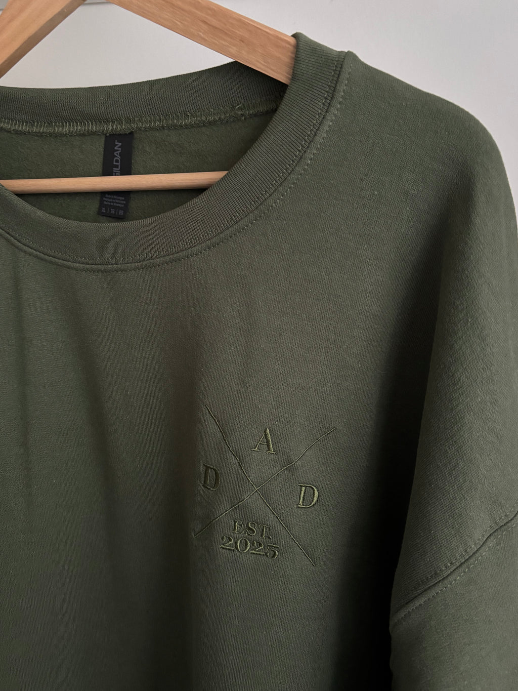 Ein Sweater in der Farbe Military Green mit einem Koordinaten Kreuz auf der linken Brust gestickt. Im Kreuz steht Dad est. 2025. Auf dem Ärmel ist der Kindername gestickt.