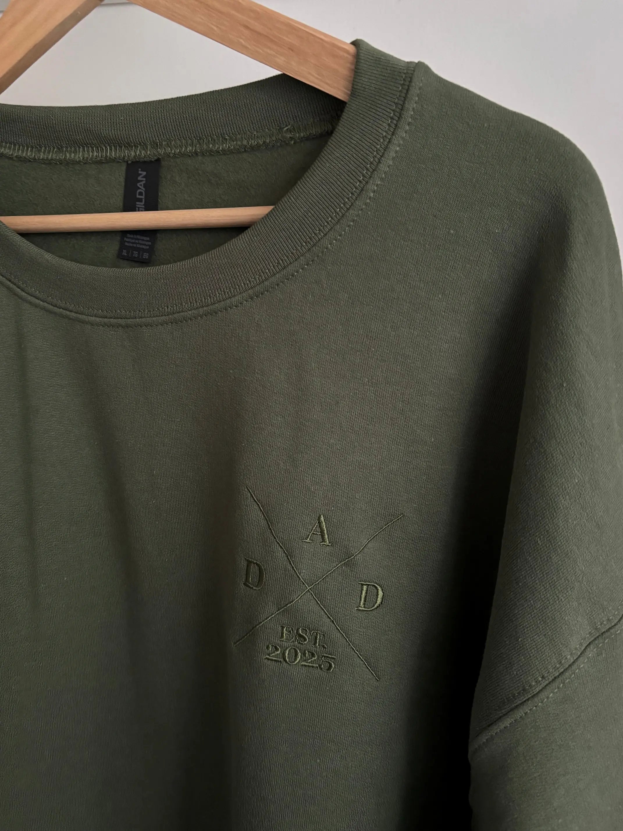 Ein Sweater in der Farbe Military Green mit einem Koordinaten Kreuz auf der linken Brust gestickt. Im Kreuz steht Dad est. 2025. Auf dem Ärmel ist der Kindername gestickt.