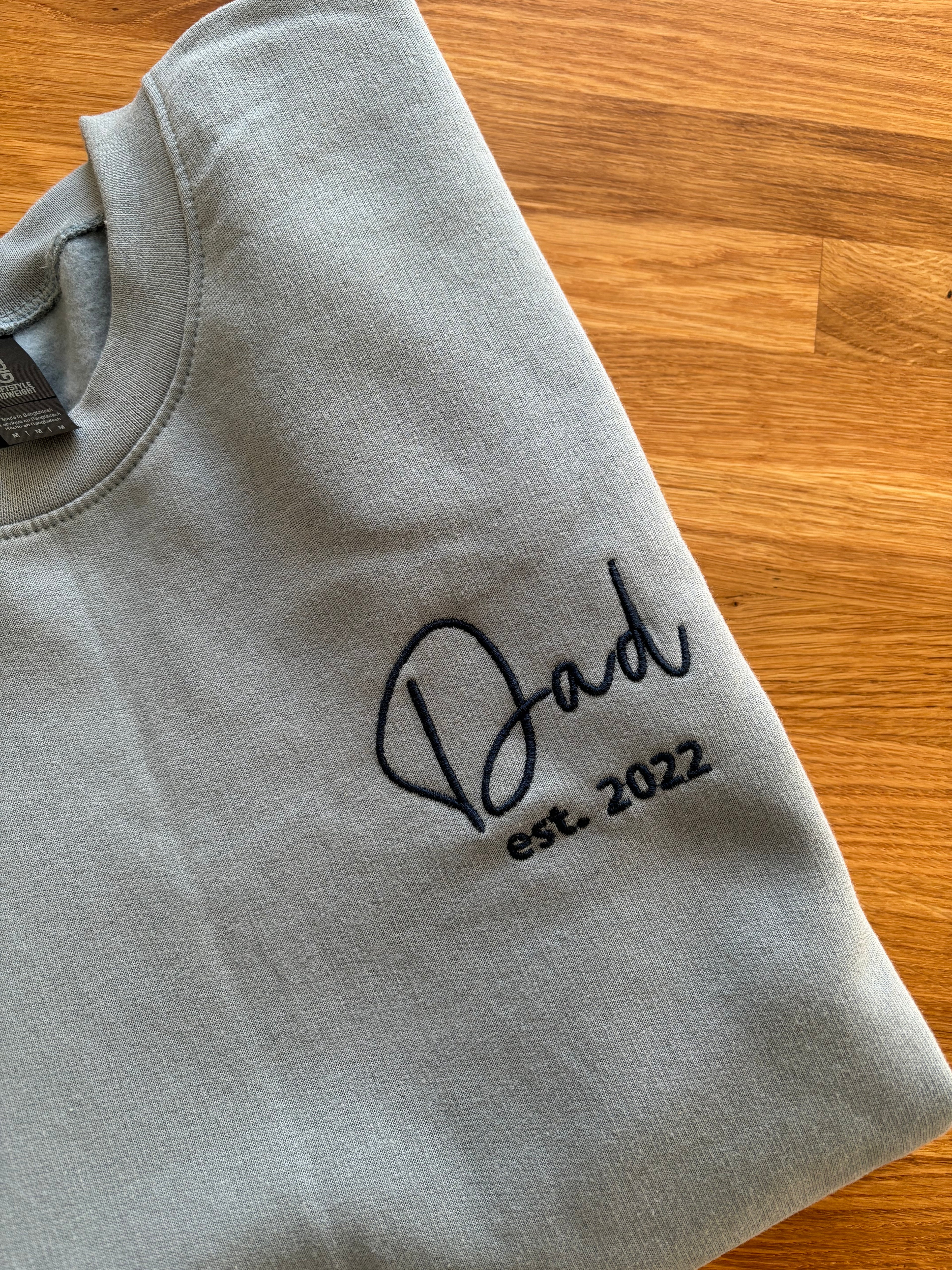Ein Sweater in der Farbe Stone Blue mit einem personalisierten Text "Dad est. 2022".