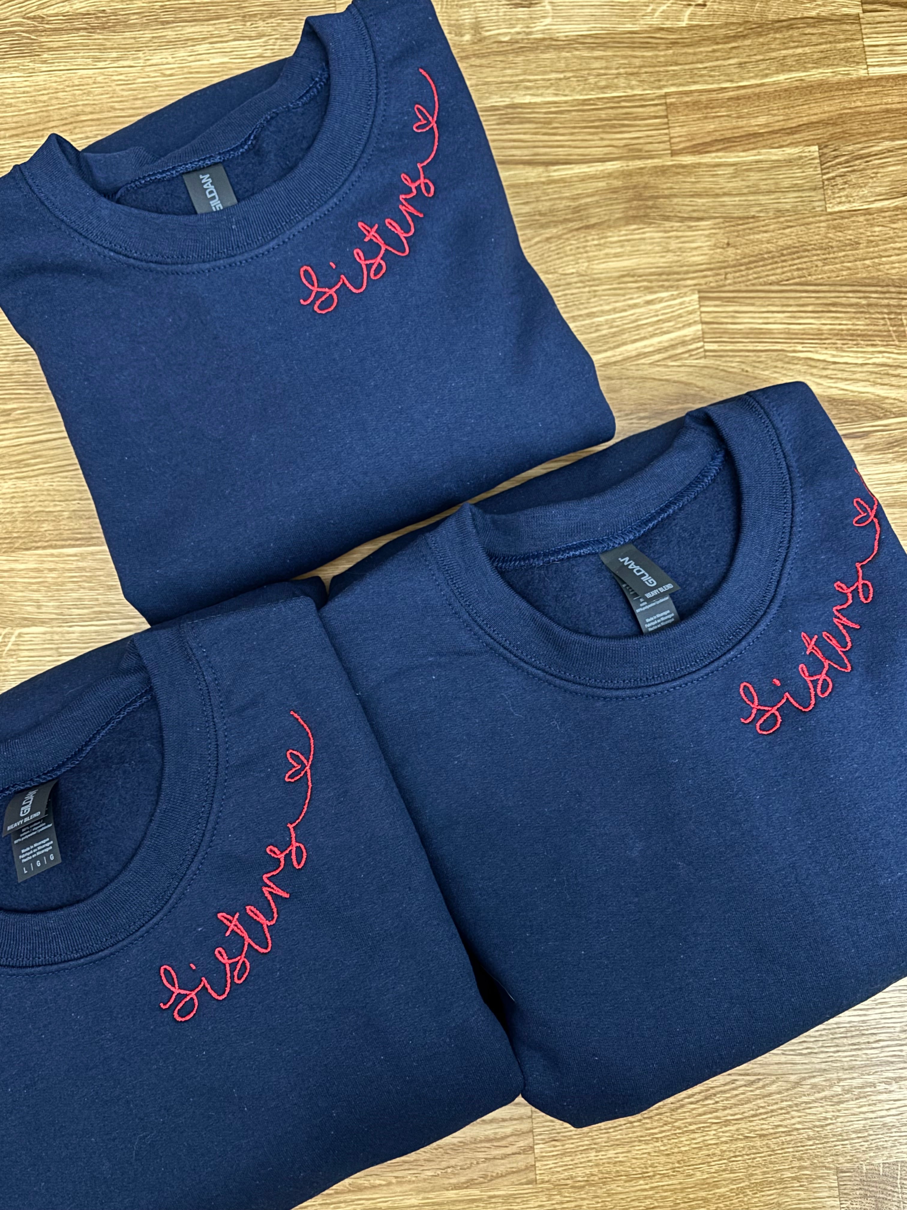 Drei Sweater in der Farbe Navy Blue mit einer roten Stickerei am Kragen: Sisters.