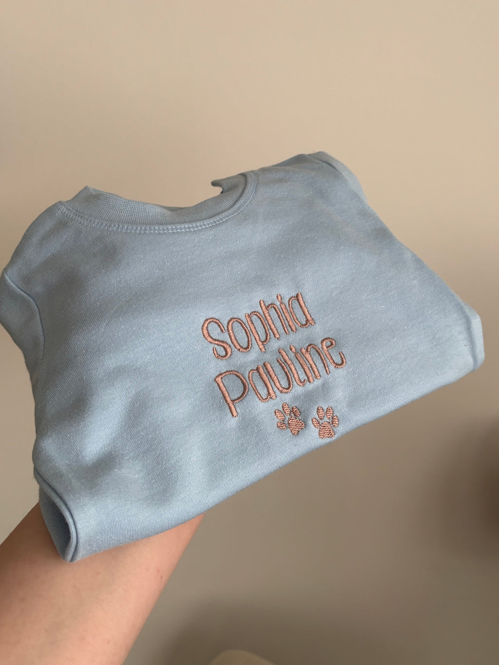 Ein Sweater für Babys in der Farbe Light Blue. Der Sweater wurde individuell mit dem Wunschtext bestickt. Der Kindername Sophoa Pauline und zwei Hundepfoten wurde in Dark beige drauf gestickt.