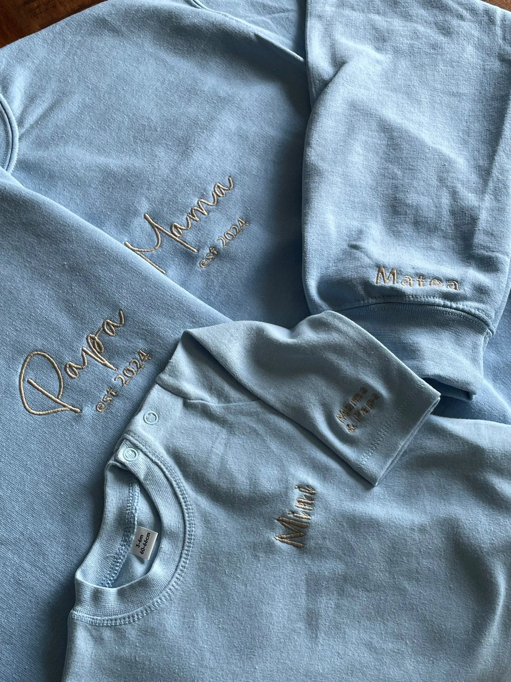 Das Sweatshirt für Babys in Kombination mit zwei Elternpullovern. Die Mutter hat einen Pullover mit Mama bestickt, der Papa hat einen Pullover mit Papa bestickt und das Baby hat ein Sweatshirt mit Mini bestickt.