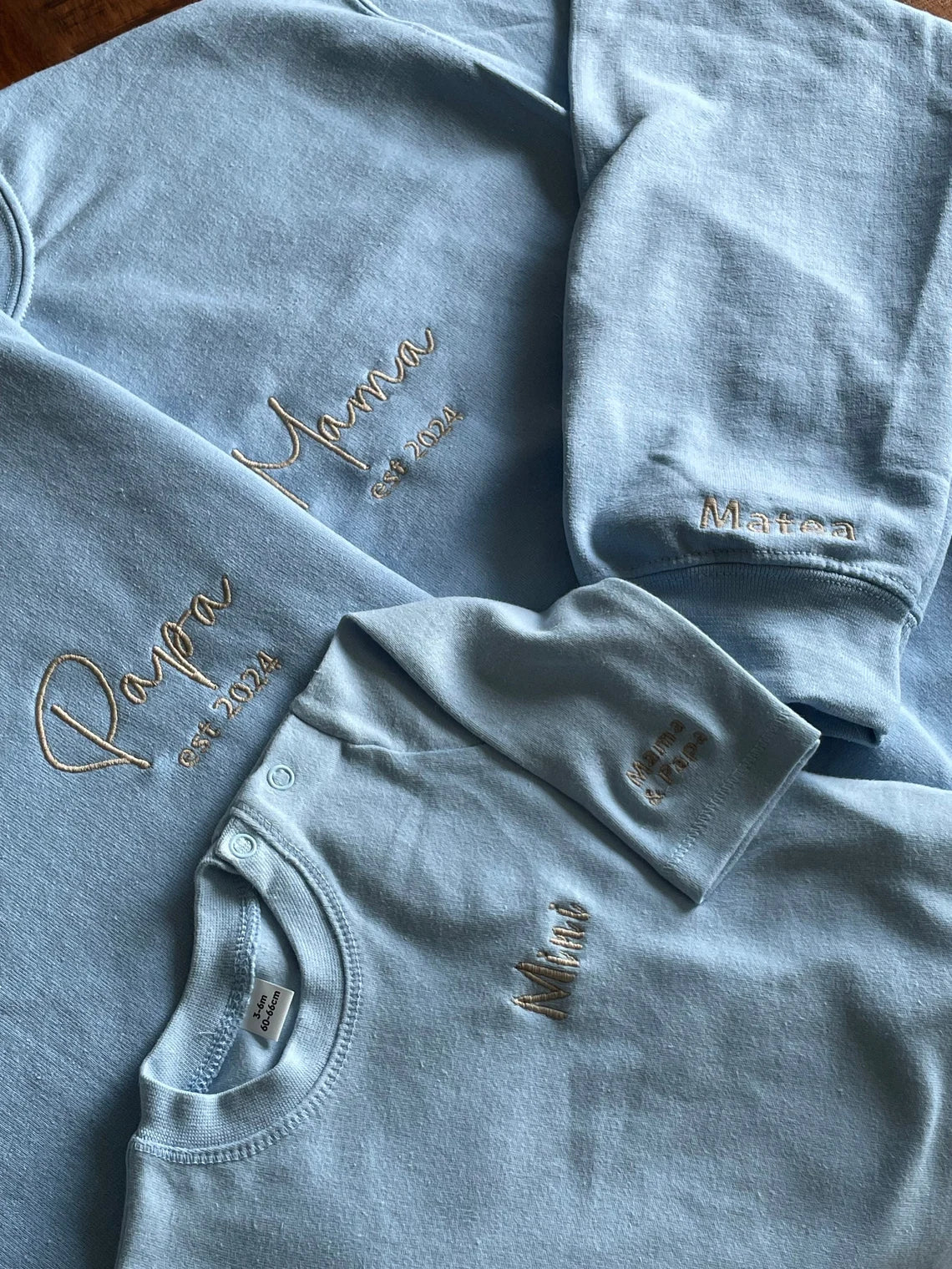 Das Sweatshirt für Babys in Kombination mit zwei Elternpullovern. Die Mutter hat einen Pullover mit Mama bestickt, der Papa hat einen Pullover mit Papa bestickt und das Baby hat ein Sweatshirt mit Mini bestickt.