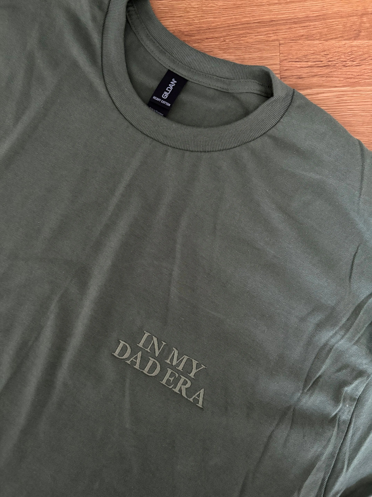 Ein T-Shirt in der Farbe Military Green mit einer Stickerei vorne links auf der Brust "In My Dad Era" in Großbuchstaben.