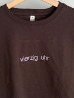 Ein braunes T-Shirt mit Rundhalsausschnitt und dem gestickten Text "vierzig uhr" in Lila. Das Shirt hängt an einem Holzbügel. Das Etikett auf dem Shirt lautet "M".