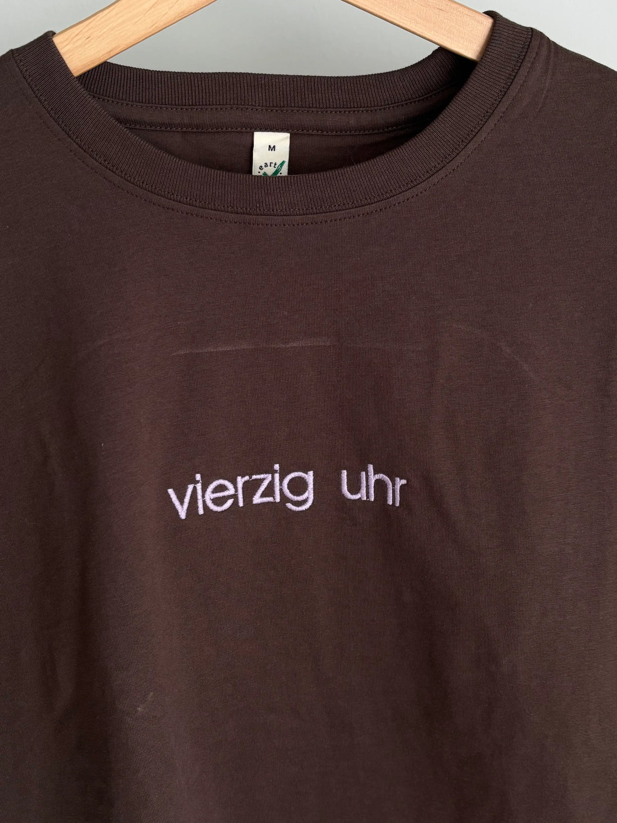 Ein braunes T-Shirt mit Rundhalsausschnitt und dem gestickten Text "vierzig uhr" in Lila. Das Shirt hängt an einem Holzbügel. Das Etikett auf dem Shirt lautet "M".