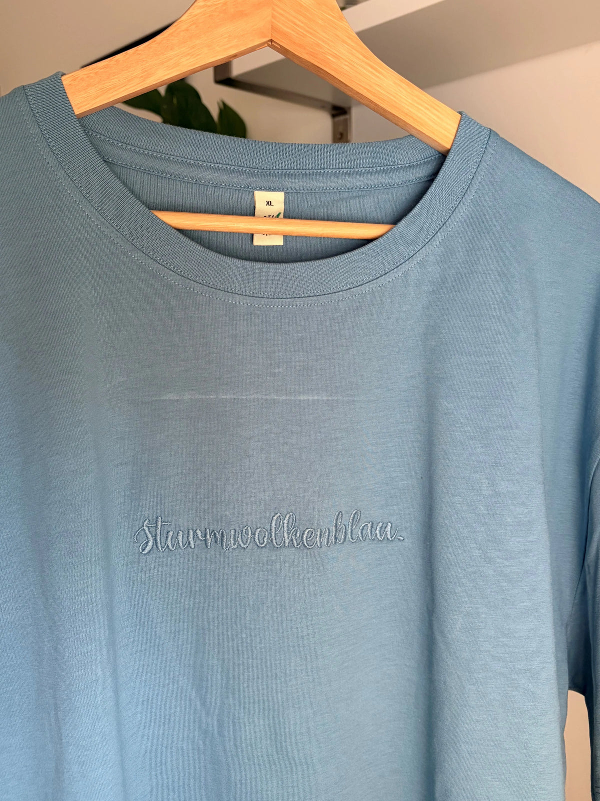 Ein T-Shirt in der Farbe Blue Dusk mit Rundhalsausschnitt und dem gestickten Text „Sturmwolkenblau“. Das Shirt hängt an einem Holzbügel. Das Etikett auf dem Shirt lautet "XL".