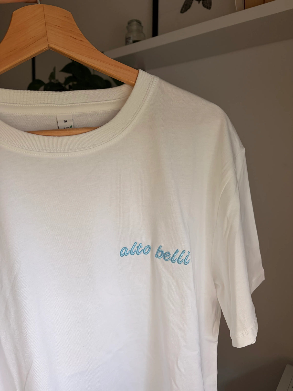 Ein weißes T-Shirt mit Rundhalsausschnitt und dem gestickten Text "alto belli“ in hellblau. Das Shirt hängt an einem Holzbügel. Das Etikett auf dem Shirt lautet "M".