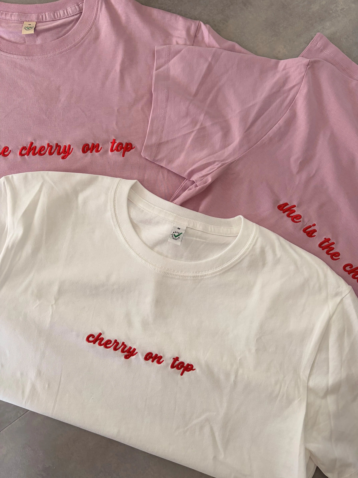 Zwei T-Shirts in der Farbe Faded Pink mit einer roten Stickerei „she is the cherry on top“. Darauf liegt ein weißes T-Shirt mit einer roten Stickerei vorne „cherry on top“.