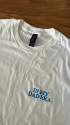 Ein weißes T-Shirt mit einer Stickerei vorne links auf der Brust "In My Dad Era" in Großbuchstaben. Das Shirt liegt auf einem Holztisch.