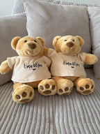 Zwei kleine Teddybären mit einem T-Shirt. Auf den Shirts ist gestickt "Oma & Opa" mit einem Herz. Der Stick ist in braun.