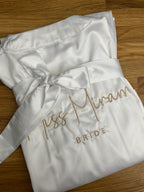 Ein weißer Morgenmantel aus Satin mit einer beigen Stickerei hinten auf dem Rücken: Miss Miram Bride.