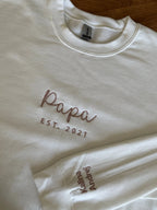 Ein Sweater in der Farbe weiß mit einer Stickerei in dark beigeauf der Brust: Papa est. 2021. Auf dem linken Ärmel sind die zwei Kindernamen gestickt: Kalina und Andrej.