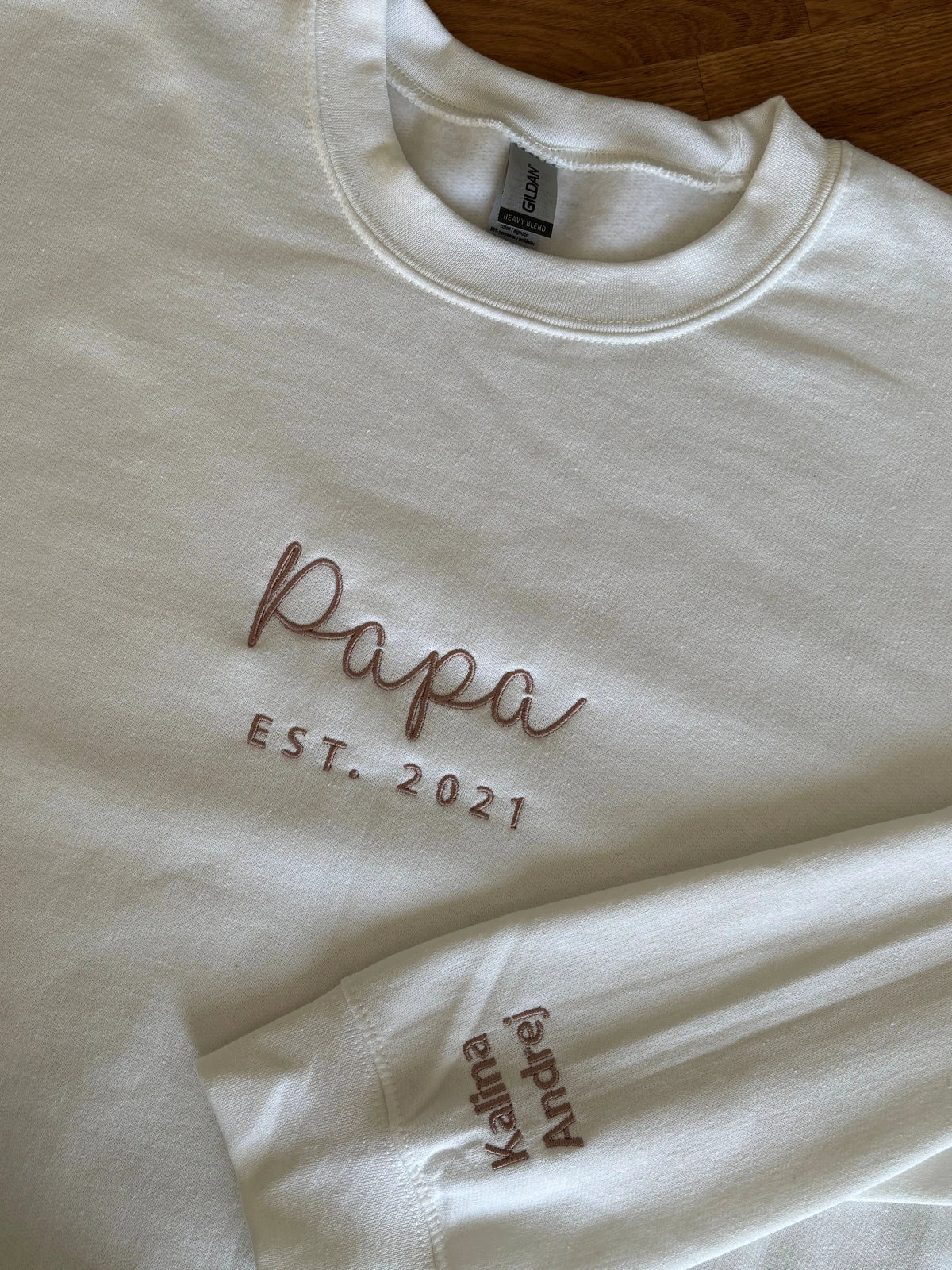 Ein Sweater in der Farbe weiß mit einer Stickerei in dark beigeauf der Brust: Papa est. 2021. Auf dem linken Ärmel sind die zwei Kindernamen gestickt: Kalina und Andrej.