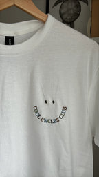Ein weißes T-Shirt mit Rundhalsausschnitt und einer Stickerei auf der linken Brust. Das Design besteht aus zwei kleinen Punkten als Augen über einer geschwungenen Linie aus mehrfarbigem Text, der COOL UNCLES CLUB bildet.