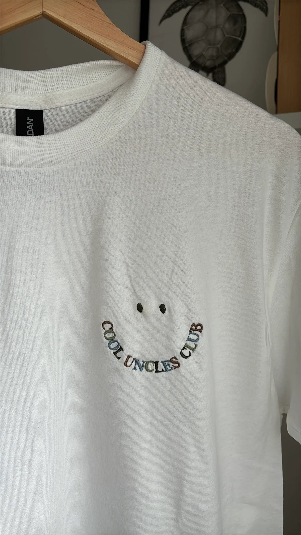Ein weißes T-Shirt mit Rundhalsausschnitt und einer Stickerei auf der linken Brust. Das Design besteht aus zwei kleinen Punkten als Augen über einer geschwungenen Linie aus mehrfarbigem Text, der COOL UNCLES CLUB bildet.