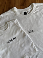 Ein T-Shirt in der Farbe Weiß mit einer schwarzen Stickerei vorne mittig auf der Brust "me". Dazu liegt ein weiteres Sweatshirt für Babys drauf mit einer schwarzen Stickerei vorne auf der Brust "mini me".