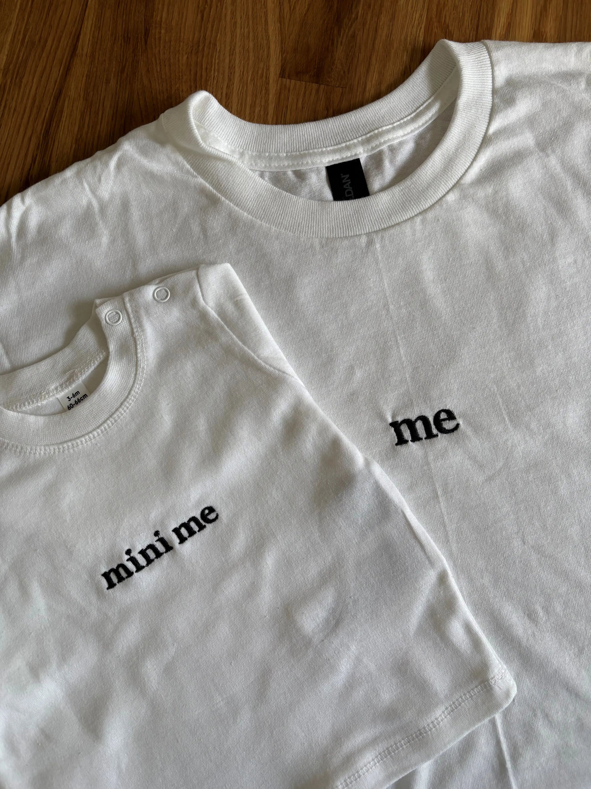Ein T-Shirt in der Farbe Weiß mit einer schwarzen Stickerei vorne mittig auf der Brust "me". Dazu liegt ein weiteres Sweatshirt für Babys drauf mit einer schwarzen Stickerei vorne auf der Brust "mini me".