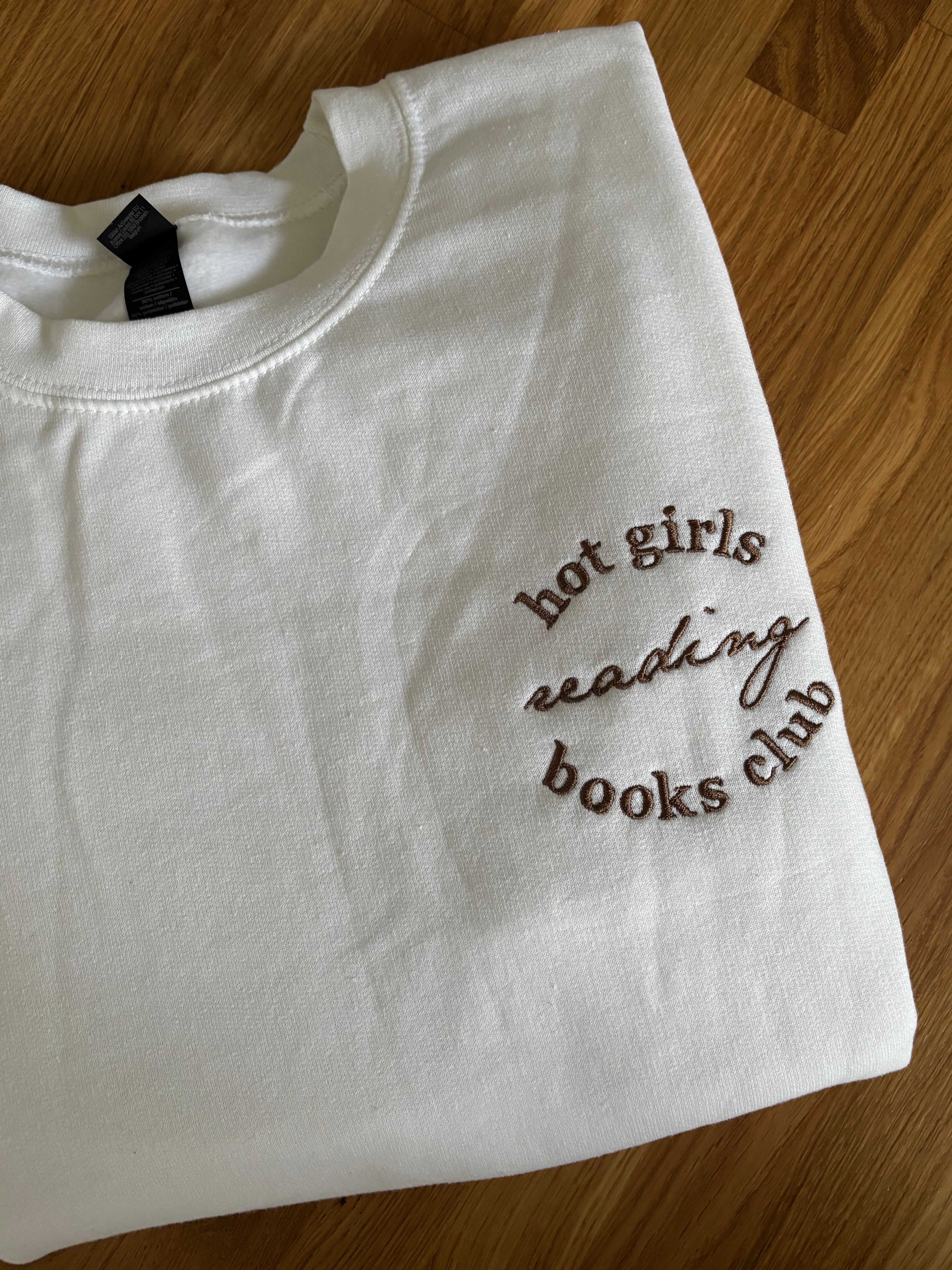 Ein weißer Sweater auf einem Holztisch. Auf der linken Brustseite ist in einem Kreis in dunkelbraun gestickt: "Hot Girls Reading Books Club". 