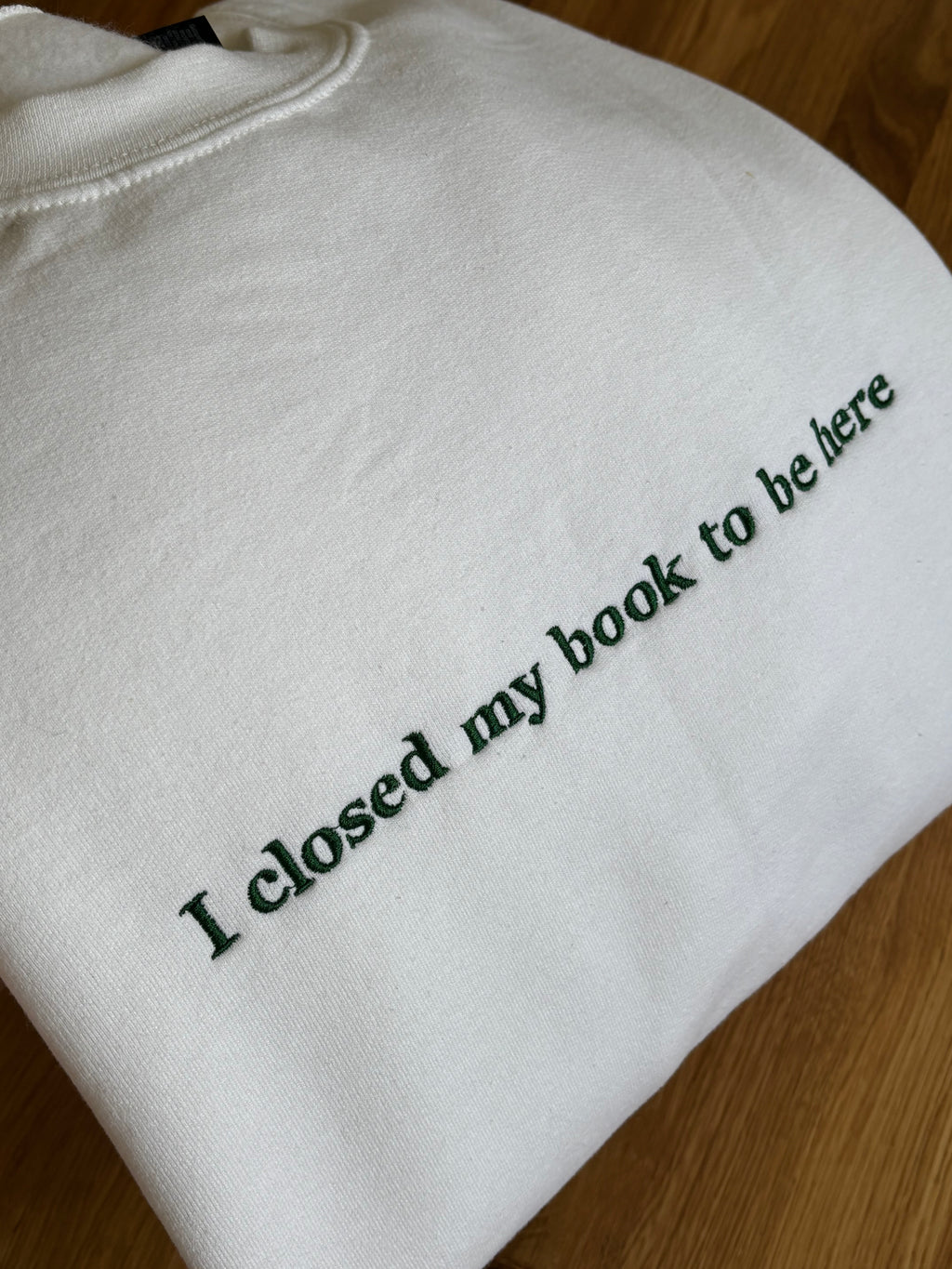 Ein weißer Sweater mit einem dunkelgrünem Stick auf der Brust "I closed my book to be here".