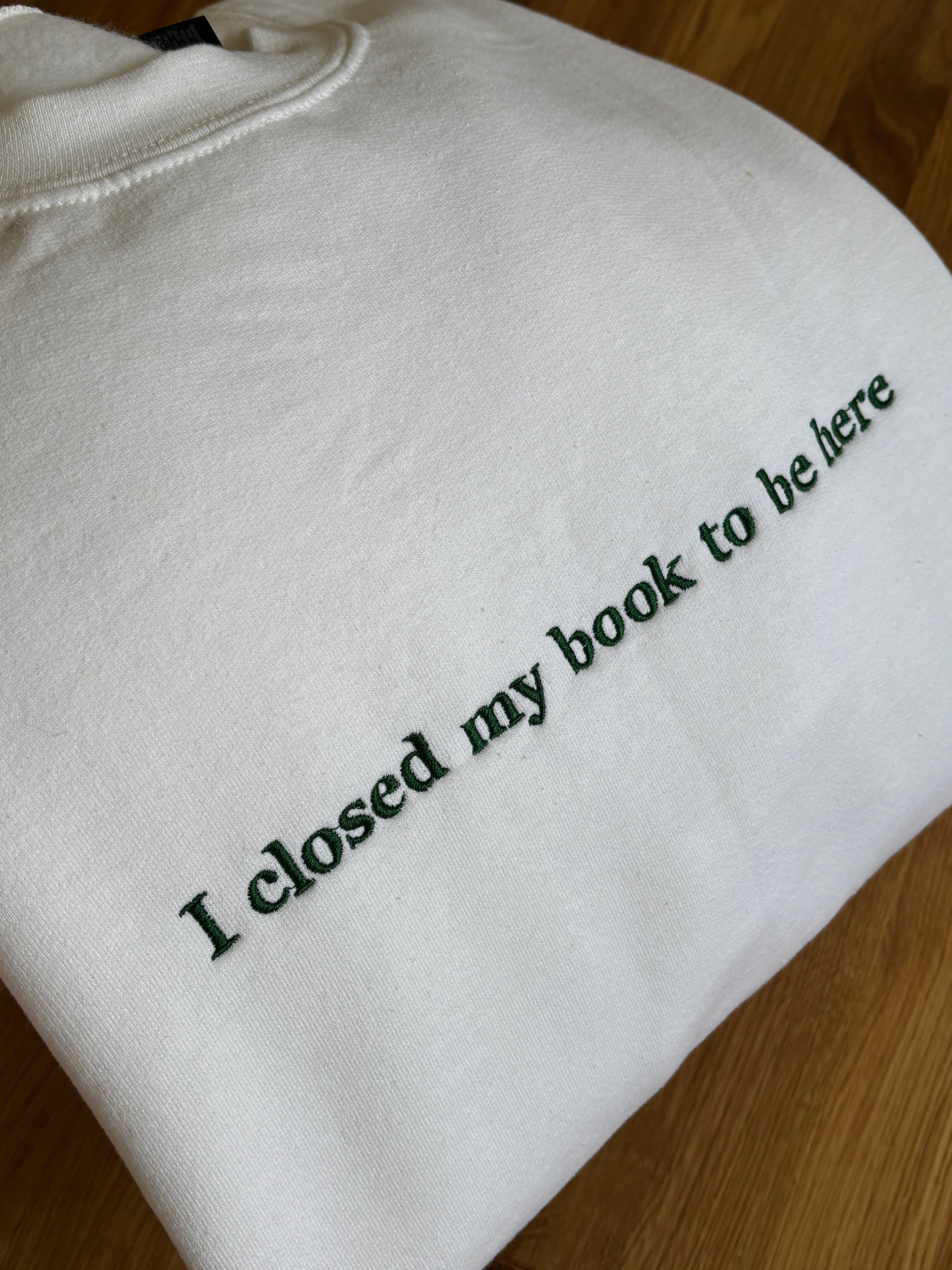 Ein weißer Sweater mit einem dunkelgrünem Stick auf der Brust "I closed my book to be here".