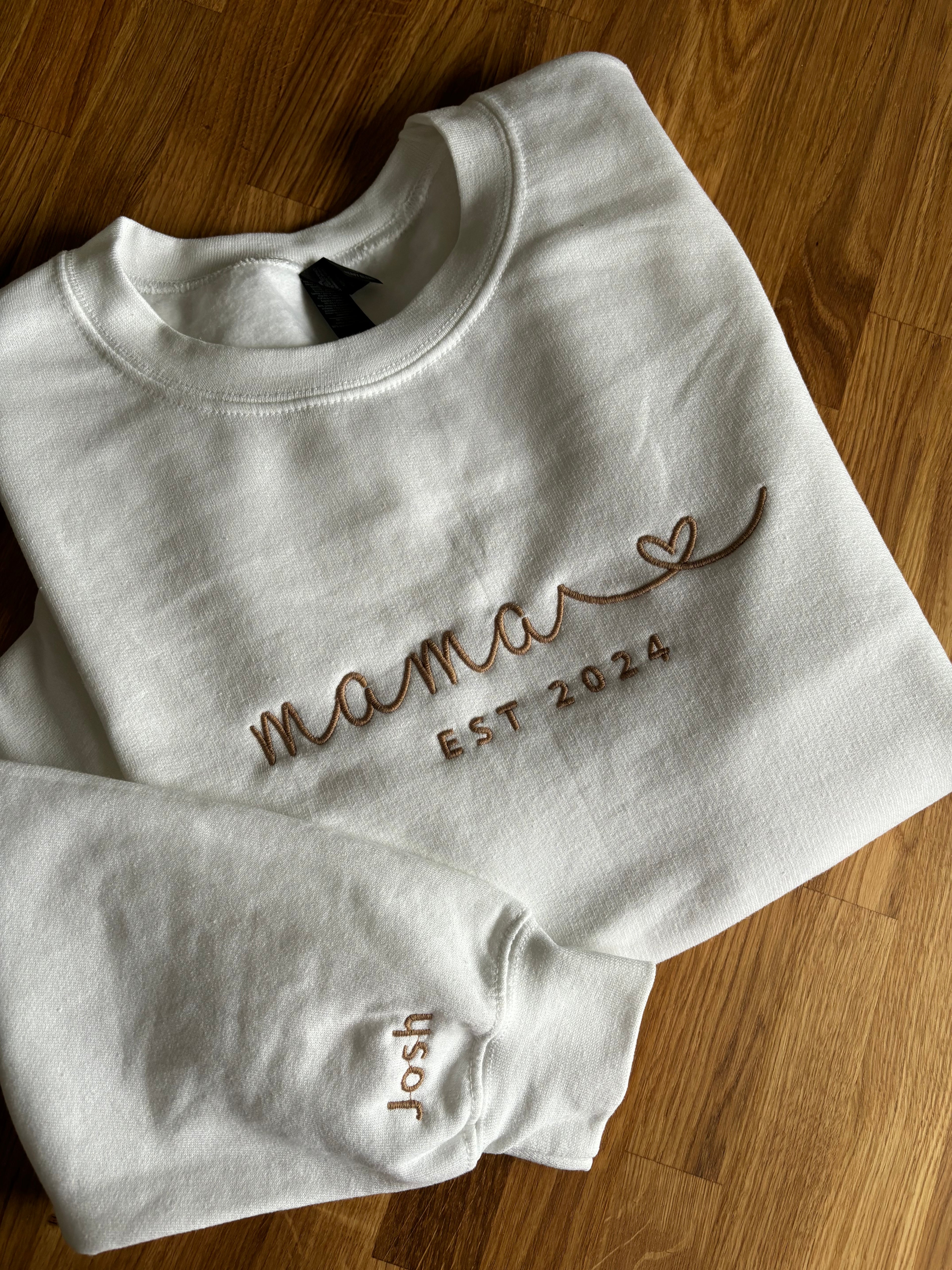 Ein weißer Sweater mit personalisiertem Stick. Auf der Brust ist gestickt "Mama Est 2024" mit einem Herz. Auf dem Ärmel ist der Name des Sohnes "Josh" gestickt.