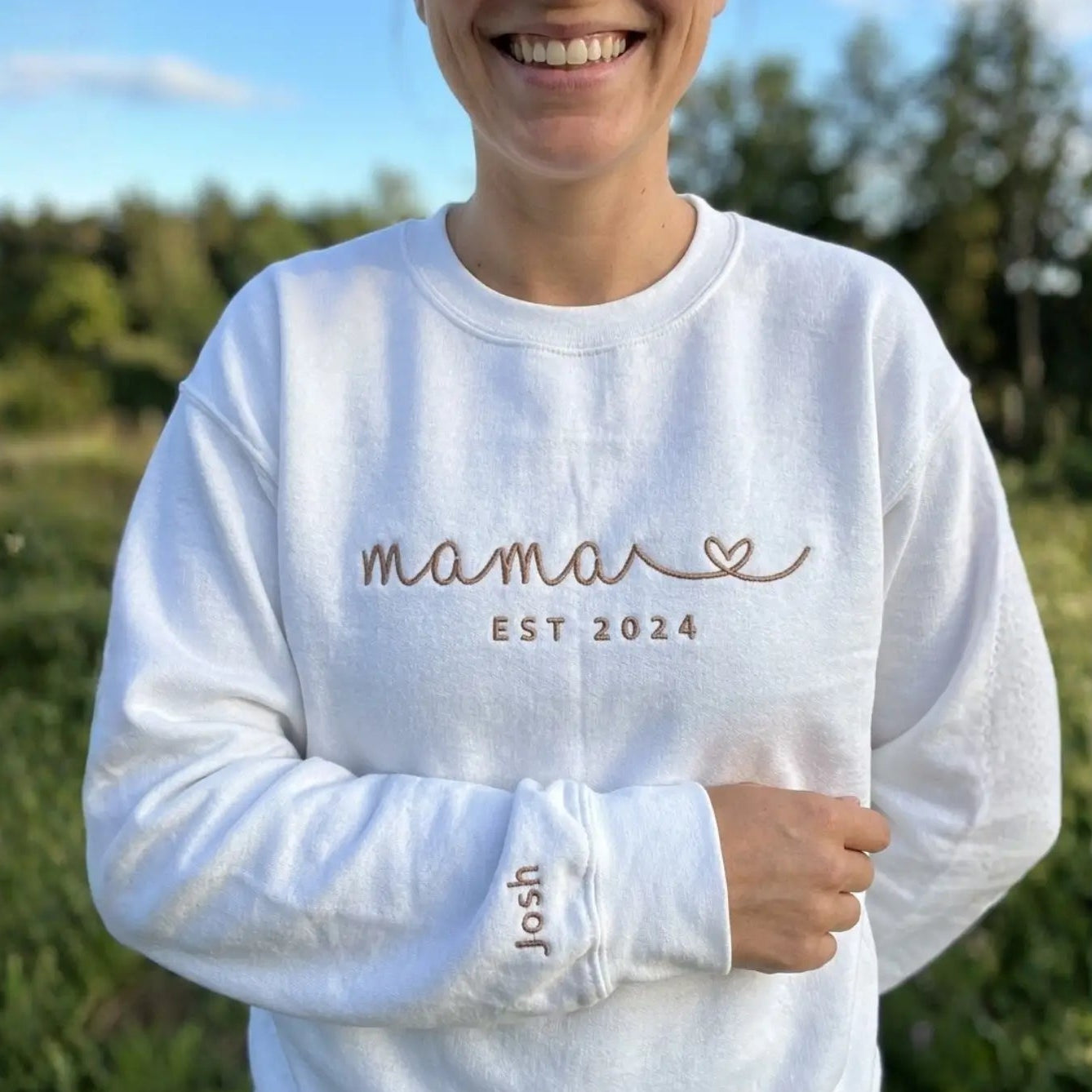 Eine Frau trägt einen personalisierten weißen Sweater mit einer Stickerei vorne: Mama Est 2024. Der Kindername ist dezent auf dem Ärmel gestickt.