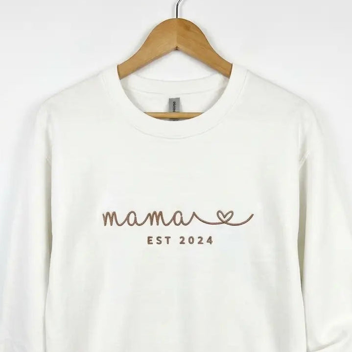 Ein weißer personalisierter Sweater mit einer Stickerei vorne Mama est 2024.