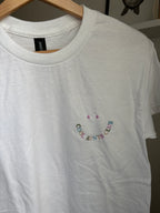 Weißes T-Shirt mit Rundhalsausschnitt. Das Shirt hat ein gesticktes Smiley-Gesicht und die Worte "COOL AUNTS CLUB" in einem geschwungenen Design. Der Text und das Smiley-Gesicht sind in verschiedenen Farben.