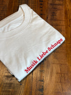 Ein T-Shirt in der Farbe Weiß mit einer roten Stickerei vorne mittig auf der Brust Musik Liebe Schnaps.