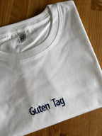 Ein weißes T-Shirt mit einer Stickerei mittig vorne auf der Brust in der Farbe Navy Blue: Guten Tag.
