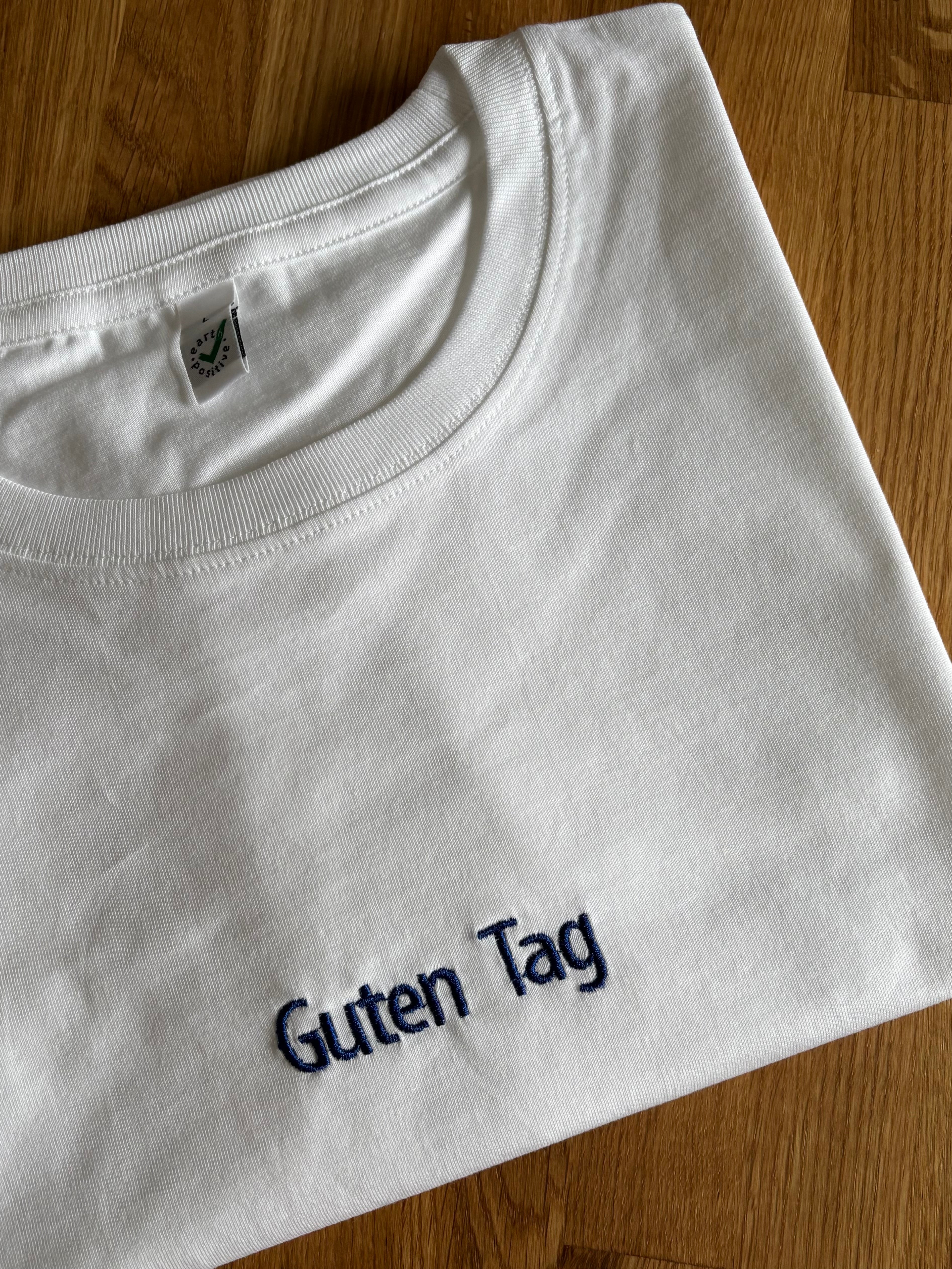 Ein weißes T-Shirt mit einer Stickerei mittig vorne auf der Brust in der Farbe Navy Blue: Guten Tag.