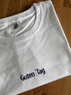 Ein weißes T-Shirt mit einer Stickerei mittig vorne auf der Brust in der Farbe Navy Blue: Guten Tag.