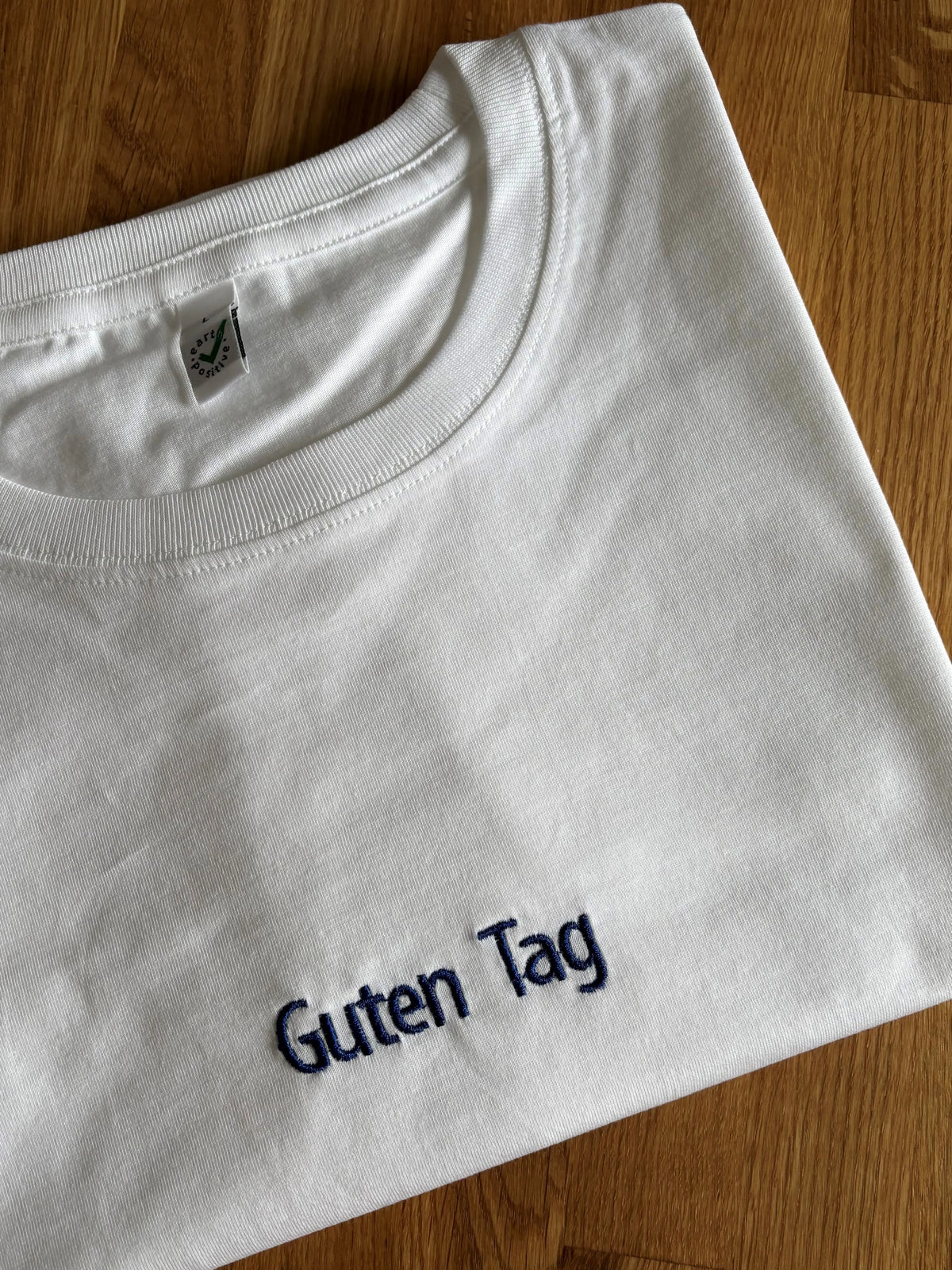 Ein weißes T-Shirt mit einer Stickerei mittig vorne auf der Brust in der Farbe Navy Blue: Guten Tag.