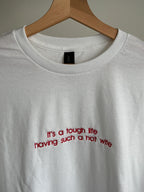 Ein T-Shirt in der Farbe Weiß mit einer roten Stickerei vorne mittig auf der Brust "It's a tough life having such a hot wife".